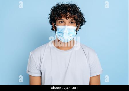 Prevention of infection. Indian or Arabian young adult curly-haired guy, with a protective medical mask on his face, looking at the camera while standing on isolated blue background Stock Photo