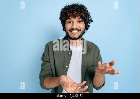 Cheerful charismatic indian or arabian guy with headset, call center worker, support service expert, standing on isolated blue background, looking at camera, smiling friendly, talking, gesturing hands Stock Photo