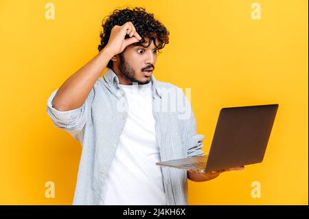 Puzzled shocked confused Indian or Arabian guy, in casual wear, take off glasses, holds an open laptop in hand, looks surprised at it, while standing on isolated orange background, get unexpected news Stock Photo