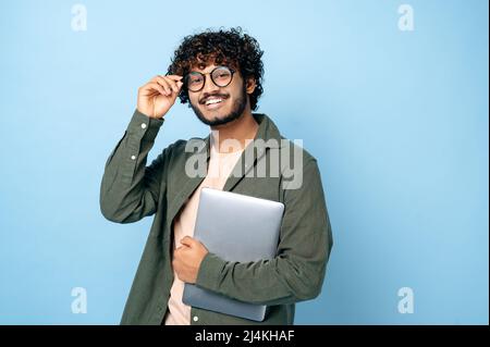 Smart handsome positive indian or arabian millennial guy, with glasses, student or freelancer, holding a laptop in hand, standing on isolated blue background, looking at the camera, smiling friendly Stock Photo