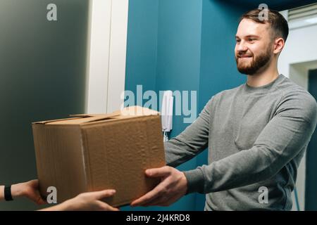 Side view of cheerful bearded courier male delivering cardboard box parcel to female customer on doorway at home. Stock Photo