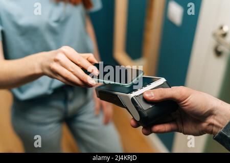 Close-up high-angle view of unrecognizable courier male giving POS wireless terminal to making contactless payment using mobile phone to young female Stock Photo