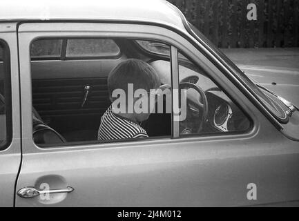 1970, historical, side view, a small boy sitting in the driver's seat of a Ford Escort car of the era holding the steering wheel, pretending to drive, England, UK. Stock Photo