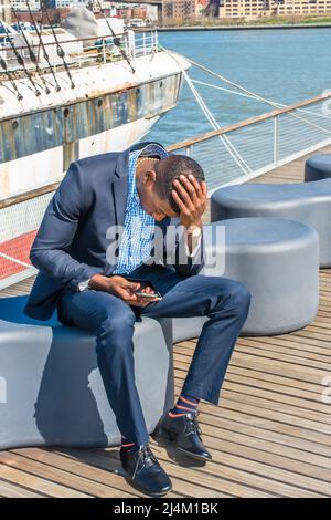 Dressing in a blue suit, leather shoes, a hand touching the top of his head , a young black businessman is sitting on the deck, looking down at his mo Stock Photo