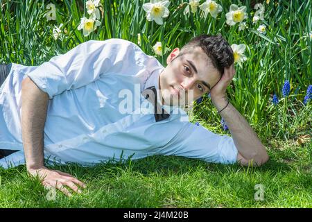 Dressing in white shirt and a black bow tie, a young handsome professional is lying  on grasses in the front of flowers to relax. Stock Photo