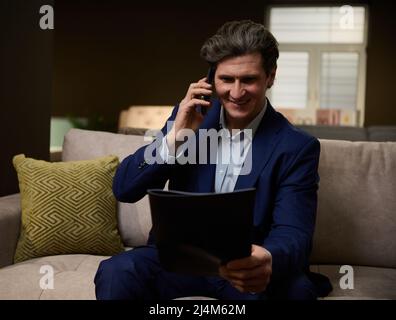 A charming successful businessman, a sales assistant in a furniture salon, holds a catalog in his hands and talks with a client on the phone, agreeing Stock Photo