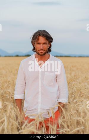 The handsome brutal young man with long brunette hairs poses in the field with wheat, suntanned man, easy beard, sunset light, white shirt, rye field Stock Photo