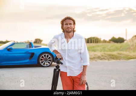 A young handsome man with long hair stands with a tripod somewhere on a country road, the photographer artist arrived in a blue sports car in the Stock Photo