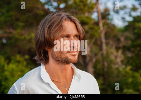 Portrait of handsome brutal man with long hair at sunset, the blue eyes, an easy bristle, he is dressed in a white shirt with a short sleeve, green Stock Photo
