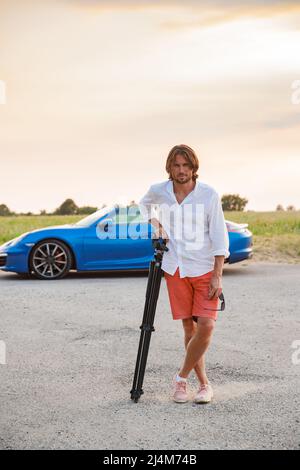A young handsome man with long hair stands with a tripod somewhere on a country road, the photographer artist arrived in a blue sports car in the Stock Photo