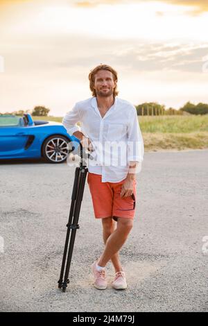 A young handsome man with long hair stands with a tripod somewhere on a country road, the photographer artist arrived in a blue sports car in the Stock Photo