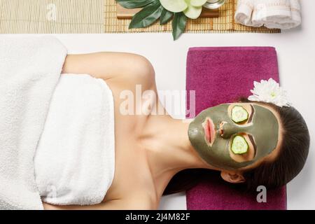 Spa Clay Mask. Woman with clay facial mask and cucumbers on eyes in beauty spa. Skincare. Beauty Concept. Close-up portrait of beautiful girl with fac Stock Photo