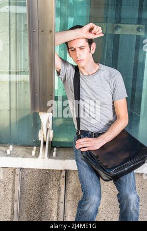 Carrying a black leather bag, dressing in a gray T shirt and blue jeans, a young guy is sitting against a metal structure, sad, depressed and seriousl Stock Photo