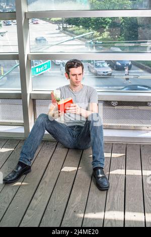 Holding a red book and a white rose, a young handsome guy is sitting on the floor by a big glass wall, reading and thinking. The background is a busy Stock Photo