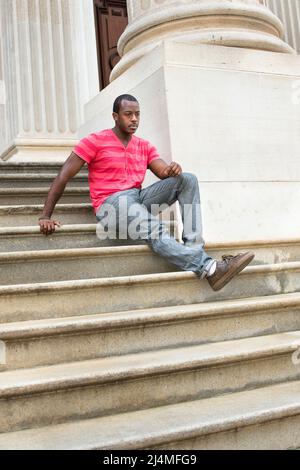 Dressing in red, pink lines T shirt,  gray pants, leather brown shoes and a bracelet, a young black guy is sitting on steps and into deeply thinking Stock Photo