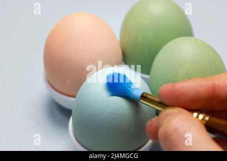 Female hand painting Easter egg with blue brush. Dyed Easter eggs in egg tray. Stock Photo