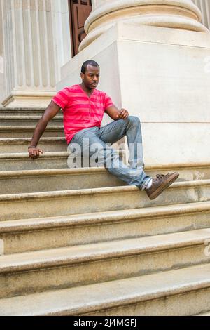 Stretching one leg, a young black guy is sitting on stairs outside an office building, relaxing and thinking Stock Photo
