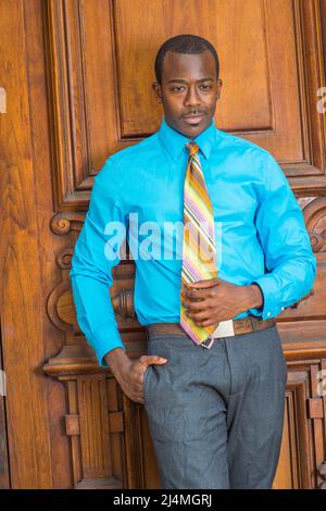 Dressing in a light blue shirt, gray pants, a pattern tie, a hand putting in pocket, a black businessman is standing by a old fashion style door, look Stock Photo