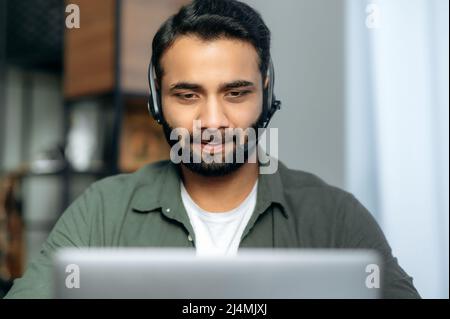 Close-up of focused serious Indian or Arabian man wearing headset, expert, consultant or mentor, looks at laptop screen, conducts online consultation. Support service concept, hotline Stock Photo