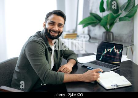 Portrait of a positive successful Indian or Arabian trader, stock market broker, investor, sitting at a desk in the office, looking at the camera, smiling friendly. Crypto charts on laptop screen Stock Photo