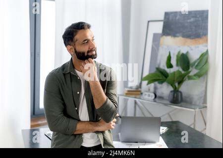 Portrait of a successful stylish Indian or Arabian businessman, manager or IT specialist, wearing casual stylish clothes, stand near his desk in the office with arms crossed, looking away and smiling Stock Photo