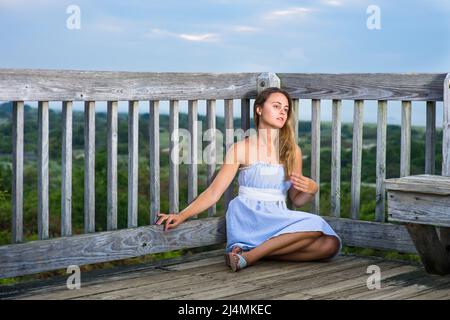 Dressing in a white blue dress, a white waistband and sandals, a pretty blonde girl is siting on a high location by the fence, thinking about you Stock Photo