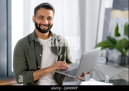 Portrait of a handsome positive Indian or Arabian successful man, programmer or freelancer in casual clothes, stands in his modern office near the table, holds an open laptop, looks at camera, smiling Stock Photo