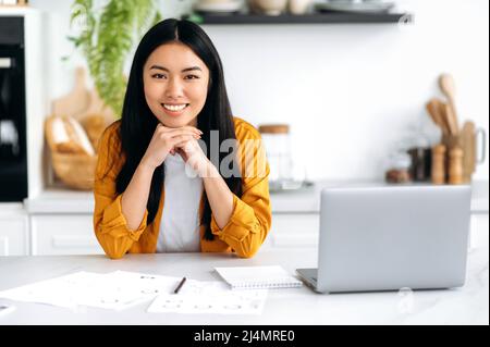 Happy pretty Asian brunette girl in stylish casual clothes, a freelancer, student or a creative designer, stands at home in the kitchen near the table and laptop, looks at camera, smiles friendly