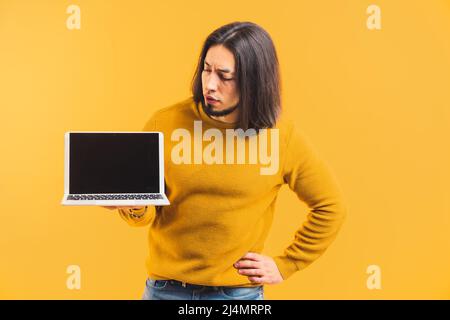 Millennial man with long dark hair holds opened laptop with a black screen, surprised look at the screen and having one hand on his waist advertisement concept isolated studio shot medium shot copy space . High quality photo Stock Photo