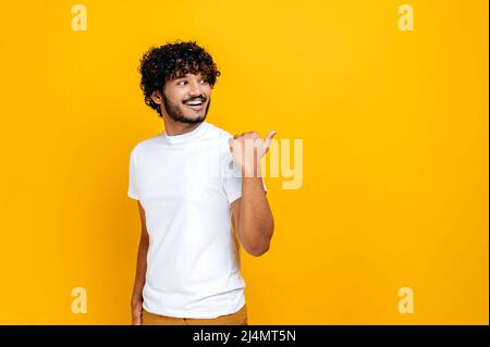 Positive curly haired indian or arabian millennial guy, looking and pointing finger to the side to an empty mock-up space, while standing over isolated orange background, smiling happily Stock Photo