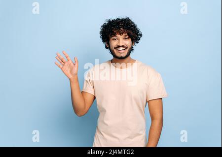 Handsome positive arabian or indian curly-haired guy, wearing casual t-shirt, standing on isolated blue background, looking at the camera, waving hand, welcome sign, smiling friendly Stock Photo