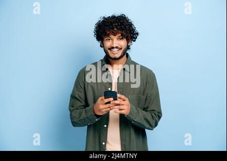 Handsome positive millennial indian or arabian guy casual wear, uses smartphone, messaging with friends online, browses social networks, news, looks at camera, smile, stand on isolated blue background Stock Photo