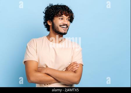 Handsome positive arabian or indian curly-haired millennial guy in a t-shirt, standing over isolated blue background with crossed arms, looking away, smiling happily Stock Photo