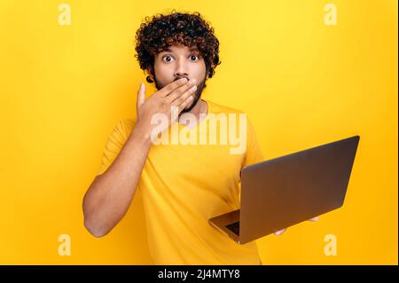 Shocked indian or arabian curly-haired guy, holding an open laptop in his hand, looks surprised at the camera, covering mouth with hand, while standing on isolated orange background Stock Photo