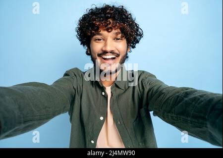 Cheerful Indian funny curly-haired guy with gladden face expression, in casual shirt, doing selfie shot on smartphone and smiling into the camera, stands on isolated blue background Stock Photo