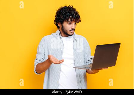 Discouraged confused indian or arabian millennial guy in casual clothes, holds an open laptop in hand, looks puzzled at it, gesturing hand, stands on an isolated orange background Stock Photo