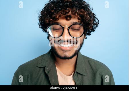 Close-up face of a curly charismatic indian or arabian guy with glasses, fooling around in front of the camera with his eyes closed, standing on an isolated blue background in a casual shirt, smiling Stock Photo
