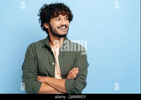 Handsome charismatic successful indian or arabian curly-haired guy in stylish clothes, standing with crossed arms over isolated blue background, looking away, smiling positively Stock Photo