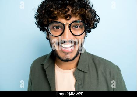 Close-up face of a curly-haired charismatic handsome indian or arabian guy with glasses, standing on an isolated blue background in a casual shirt, smiling happily Stock Photo