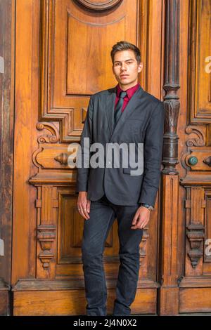 Dressing in a red undershirt, a black blazer,  jeans,  tie,  a handsome college student is standing in the front o old fashion style door, looking at Stock Photo