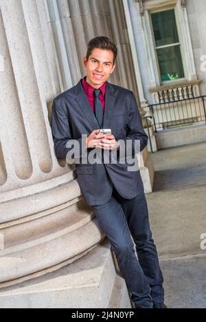 Dressing in a red undershirt, a black blazer,  jeans,  tie, a young handsome college student is leaning against a column outside an office, checking m Stock Photo