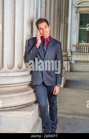 Dressing in a red undershirt, a black blazer,  jeans,  tie, a young handsome college student is leaning against a column outside an office, smiling, m Stock Photo