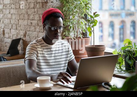 Pensive young African American freelancer man in glasses distance working on laptop in cafe Stock Photo