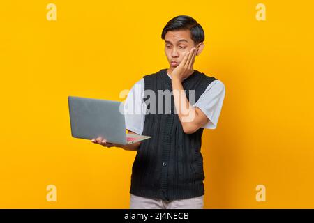 Portrait of smiling young Asian man holding laptop with hand on cheek on yellow background Stock Photo
