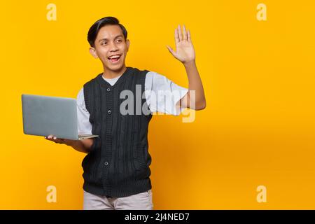 Portrait of smiling young Asian man holding laptop and gesturing wave hand on yellow background Stock Photo