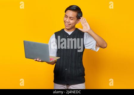 Portrait of cheerful Asian young man holding laptop and waving hand on yellow background Stock Photo