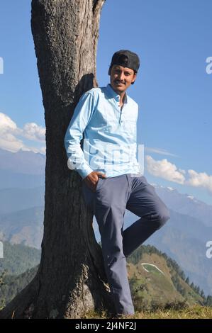 A happy young guy looking at camera while posing with leaning against tree in hilly area Stock Photo