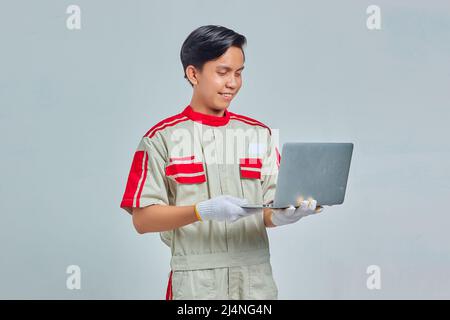 Portrait of smiling handsome mechanic man wearing uniform holding and using laptop on gray background Stock Photo