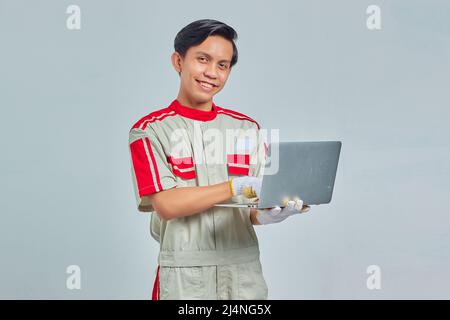 Portrait of smiling handsome mechanic man wearing uniform holding and using laptop on gray background Stock Photo