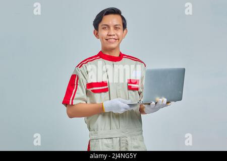 Smiling handsome young mechanic man wearing uniform holding laptop and looking at camera isolated on gray background Stock Photo